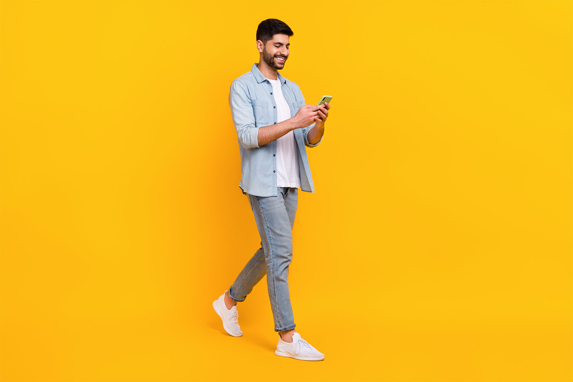 A cheerful young man in casual attire walking and looking at his smartphone against a bright yellow background.